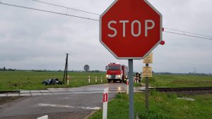Zdjęcie przedstawiające znak drogowy ,,Stop&quot; na przejeździe  kolejowym, w tle widoczny rozbity samochód i auto policji i strazy pożarnej