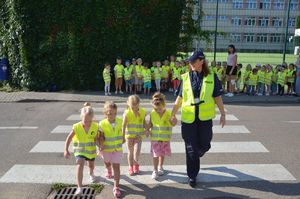 Fotografia kolorowa: dzieci w wieku przedszkolnym przy przejściu dla pieszych z policjantką.