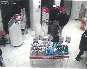 Fotografia kolorowa: dot. kradzieży sklepowej, widzimy wnętrze drogerii - półki z towarem oraz dwóch mężczyzn w czarnych kurtkach i czapkach z daszkiem (czarna i biała).