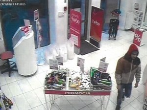 Fotografia kolorowa: dot. kradzieży sklepowej, widzimy wnętrze drogerii - półki z towarem oraz mężczyzną w brązowej kurtce z czerwonym kapturem na głowie.