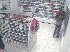 Fotografia kolorowa: dot. kradzieży sklepowej, widzimy wnętrze drogerii - półki z towarem oraz dwóch mężczyzn: jeden w czarnej kurtce a drugi w brązowej z czerwonym kapturem na głowie.