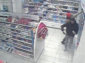 Fotografia kolorowa: dot. kradzieży sklepowej, widzimy wnętrze drogerii - półki z towarem oraz dwóch mężczyzn: jeden w czarnej kurtce a drugi w brązowej z czerwonym kapturem na głowie.