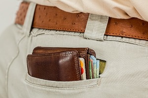 Brązowy portfel z którego wystaje gotówka i karty płatnicze umieszczony w prawej tylnej kieszeni jasnobrązowych spodni