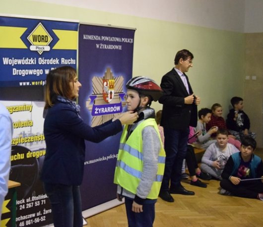 Przedstawiciel Wojewódzkiego Ośrodka Ruchu Drogowego zakłada uczniowi kask przez jazdą na torze rowerowym.